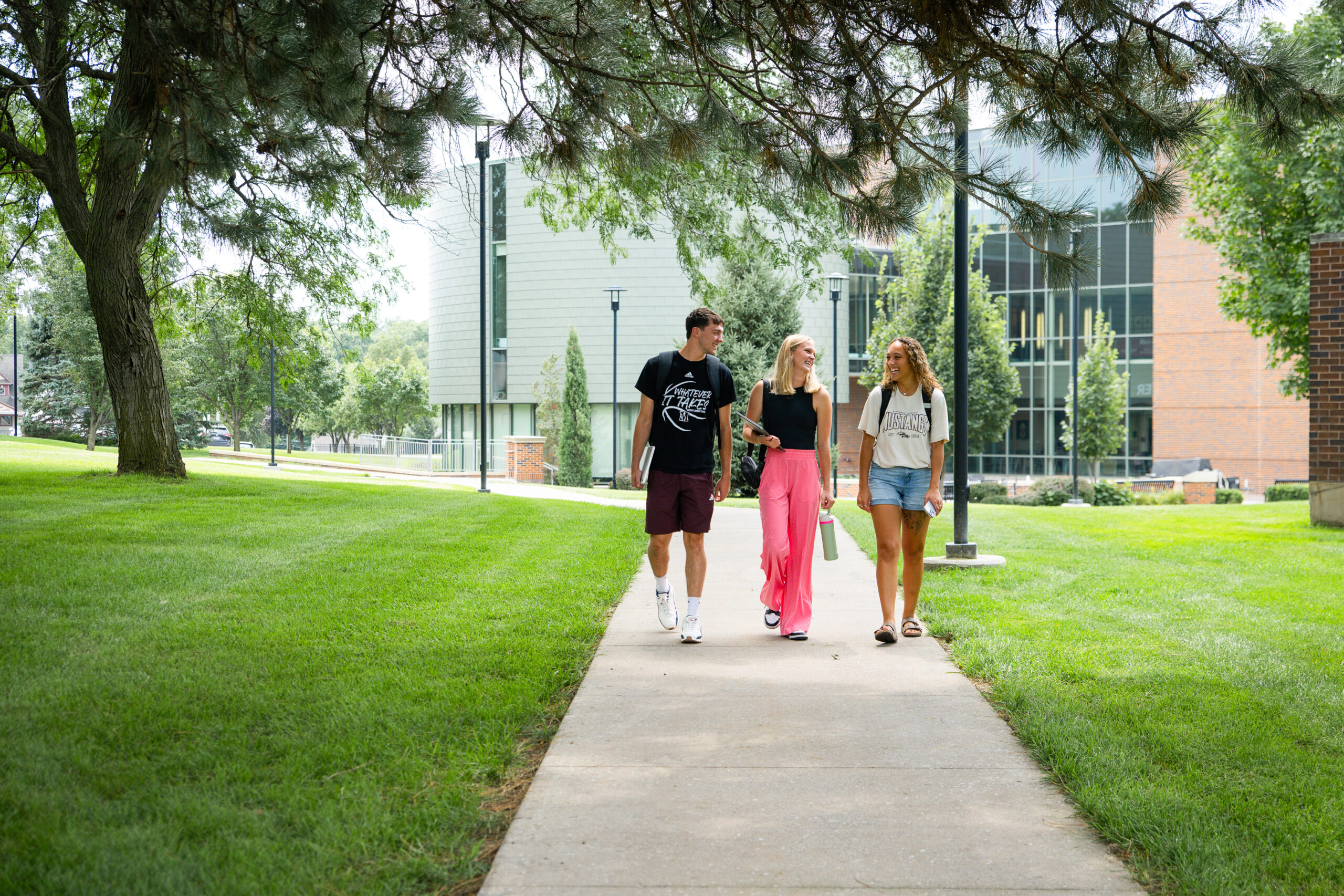 One male and two female students walking together on campus while talking and smiling. They are walking on a sidewalk path on campus surrounded by trees on both sides.