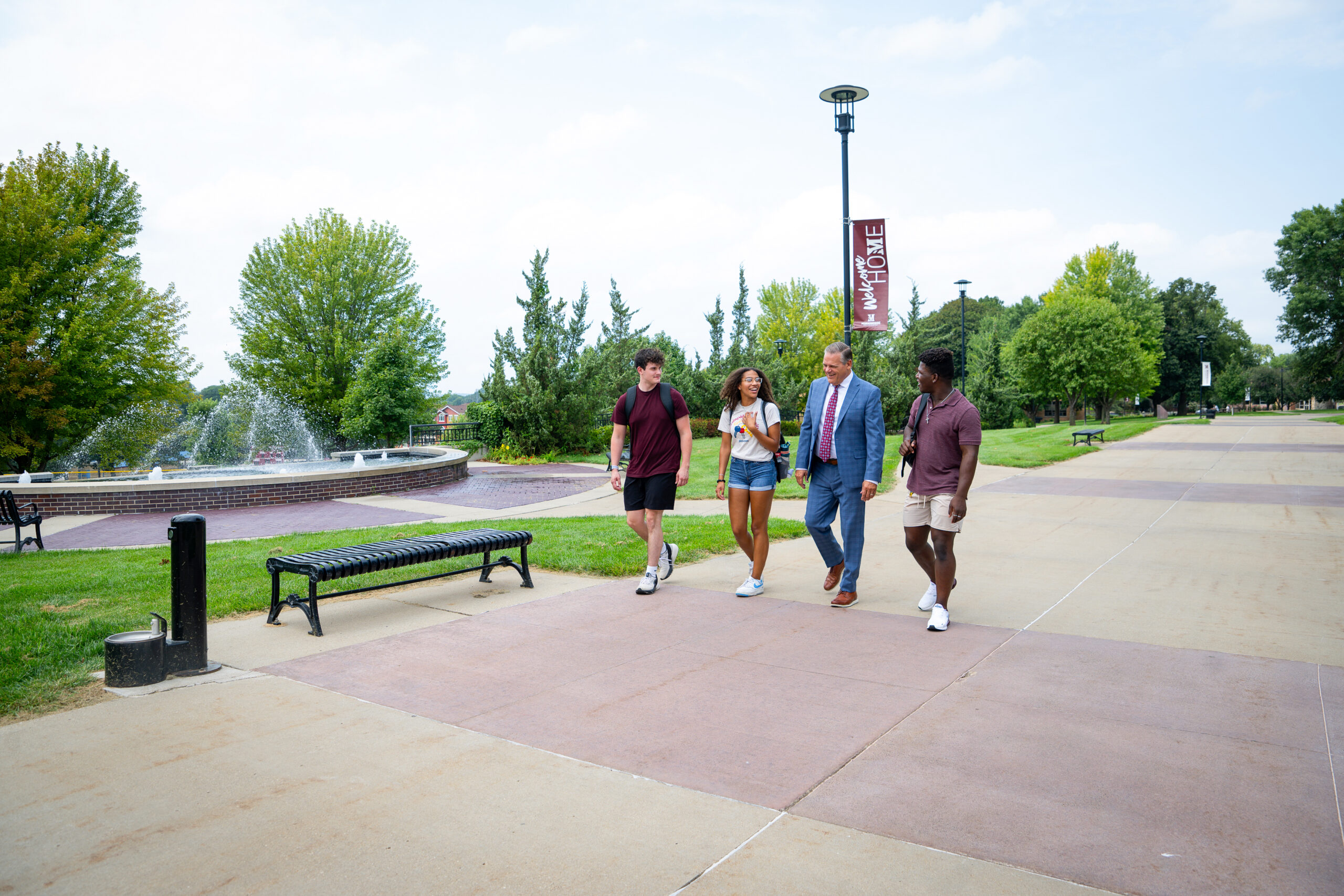 President Chad Benson walking with 3 other students on the campus mall by the water fountain feature.