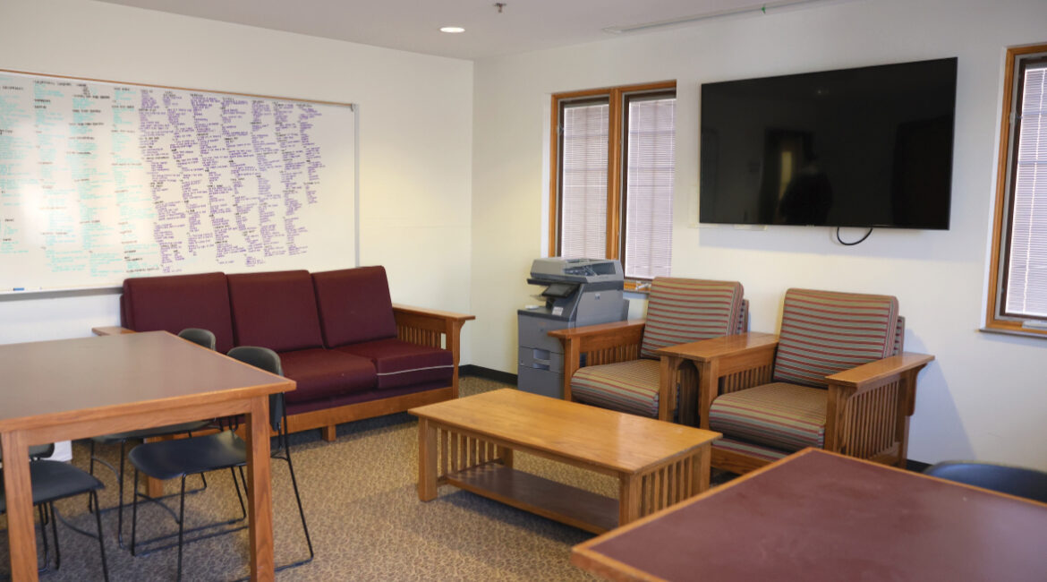 Lags lounge showing a couch, two chairs, a coffee table, and two rectangle tables with 4 chairs. A TV hangs on the wall and a printer is off the side against the wall. A giant white board with writing hangs on the wall to the left.
