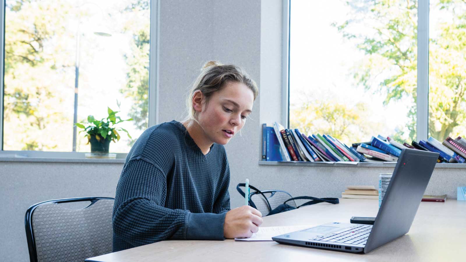 student sits at a table with computer and is writing in a notebook.