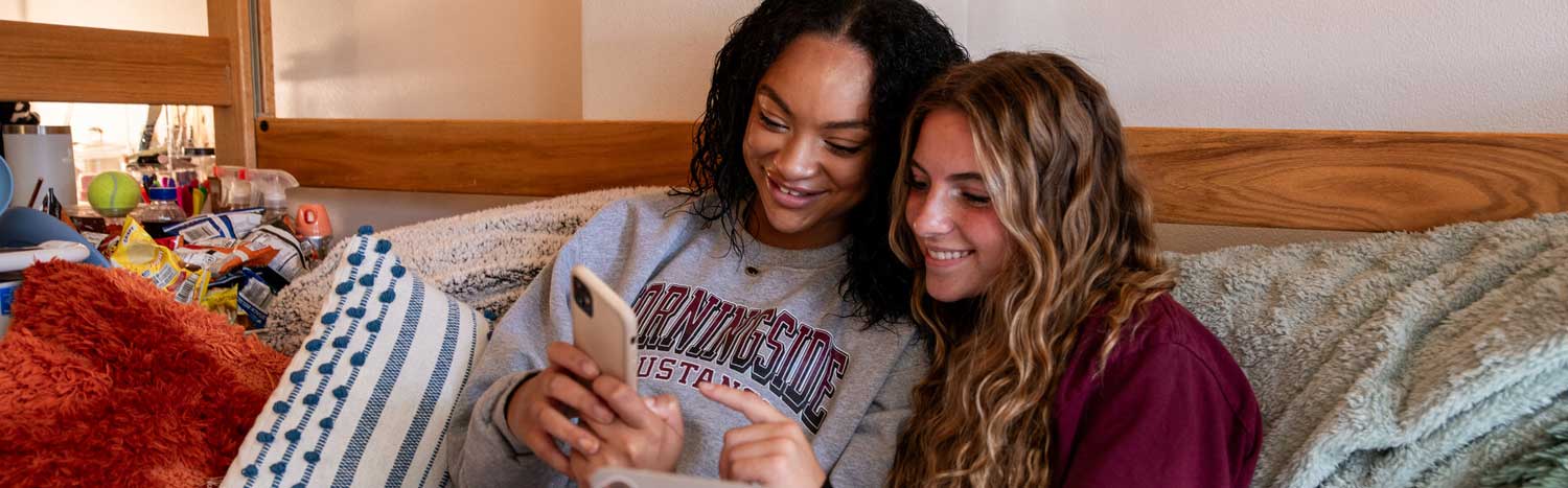 Two Students Sitting In Dorm Room With Phone