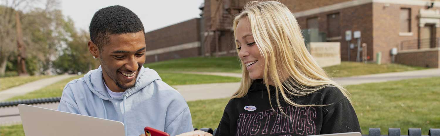 Two Students Sitting On Bench Looking At Phone