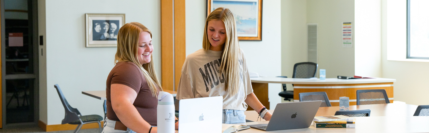 Two students standing in the library looking at their laptops