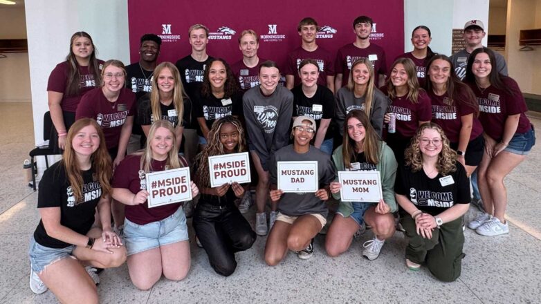 24 college student stand in front of a maroon banner holding welcome signs. 
