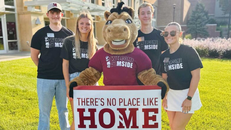 four students standing with the Morningside mascot in between in front of a sign that says There's not place like Home.