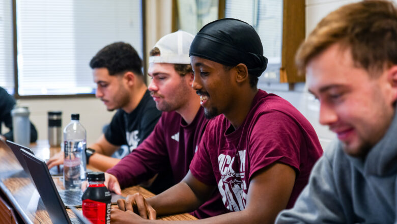 Students sitting in a row of desks in a classroom, taking notes on their laptops