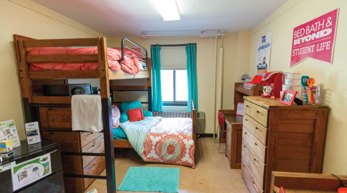 A room in Roadman Hall. One lofted bed and one floor level bed on the left . Two desks, two chairs, and a dresser on the right. Room is decorated with teal and pink. 