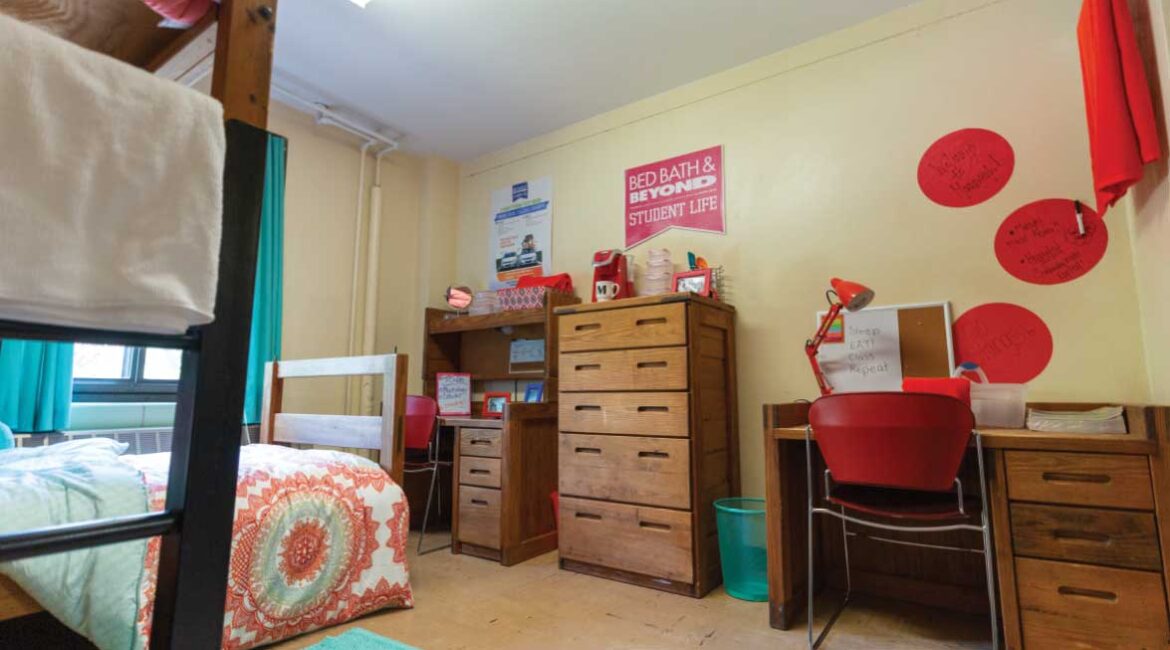 A room in Roadman Hall. One lofted bed and one floor level bed on the left . Two desks, two chairs, and a dresser on the right. Room is decorated with teal and pink. 