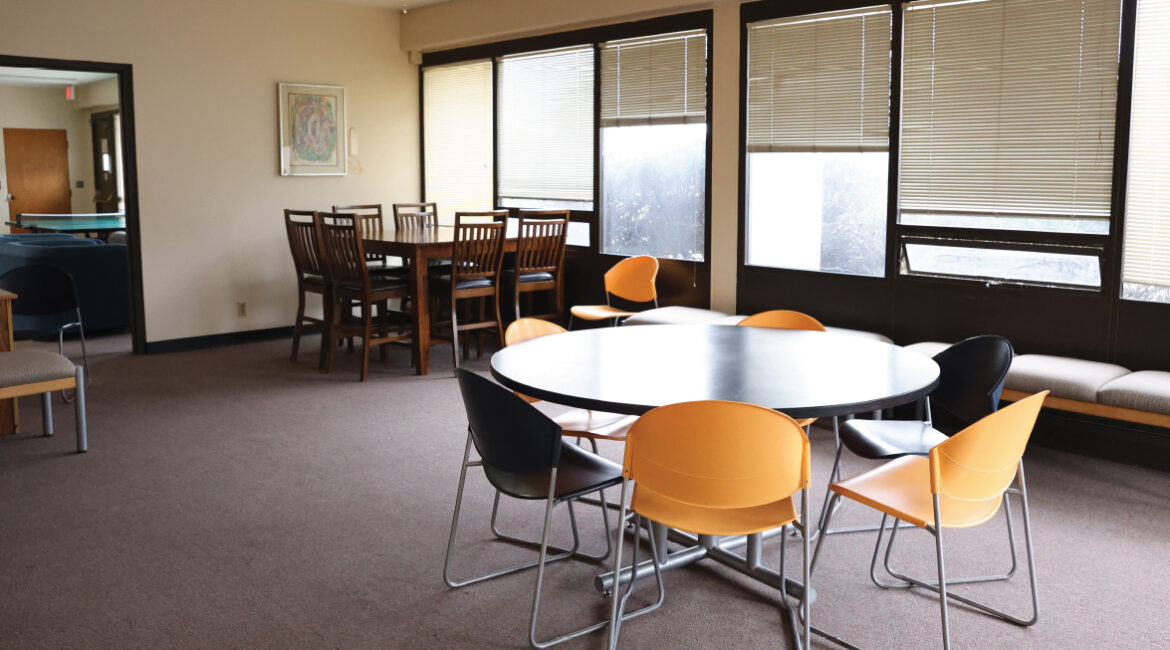 A lobby room in the plex. A wall of windows sits on right. Two tables with multiple chairs in the center of the photo. 