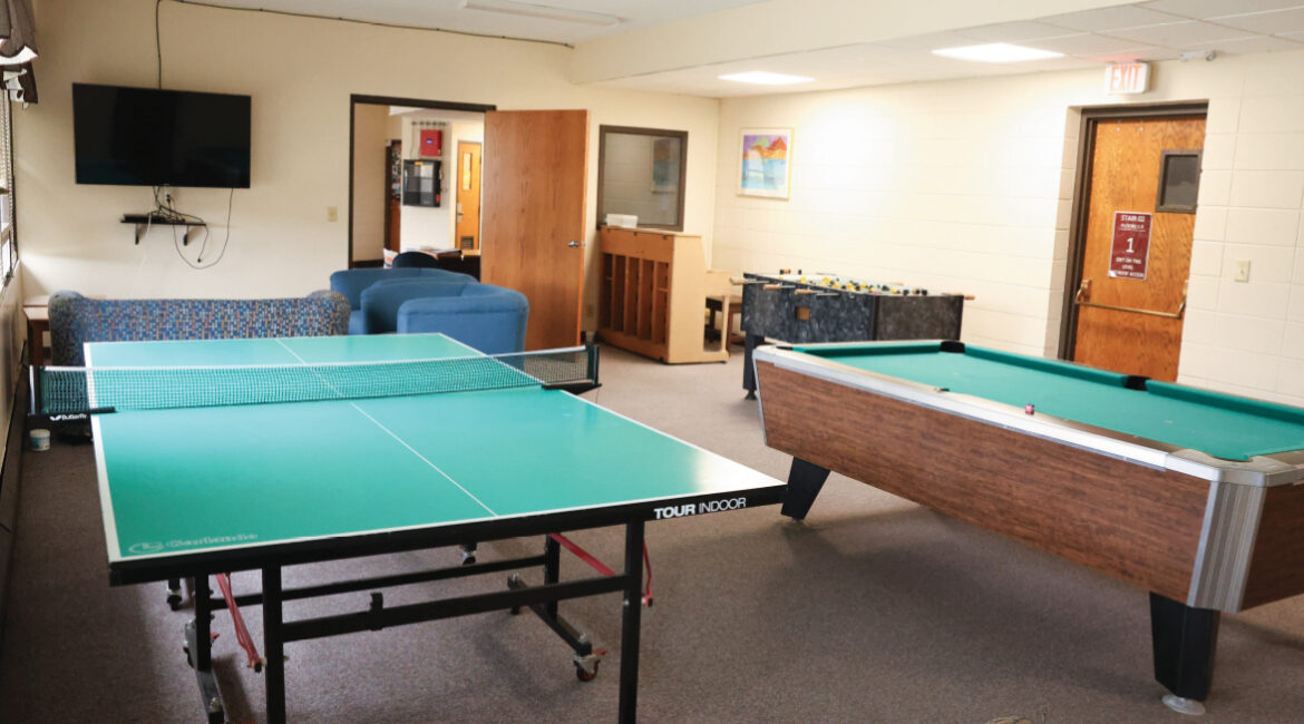 The informal lobby in the Plex. There is a wall of windows to the left. There is a ping pong table, a pool table, and a foosball table in the room. A TV with a few comfortable chairs sit in the far corner. 