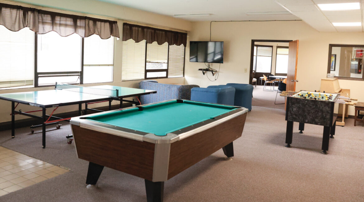 The informal lobby in the Plex. There is a wall of windows to the left. There is a ping pong table, a pool table, and a foosball table in the room. A TV with a few comfortable chairs sit in the far corner. 