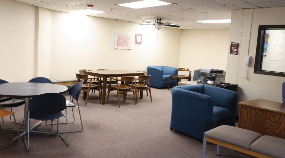 a lobby in the plex. Throughout the room are 2 tables with chairs and a few comfortable chair options. A printer sits off to the left corner.