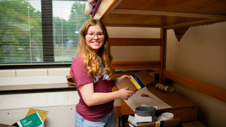 student smiling while unpacking a box in dorm room