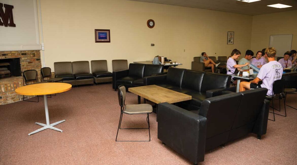 A formal lounge in Roadman Hall. Seating near a brick fireplace with the Morningside M mounted on the wall on top of the fireplace. A group of students sit at a table off to the right. 