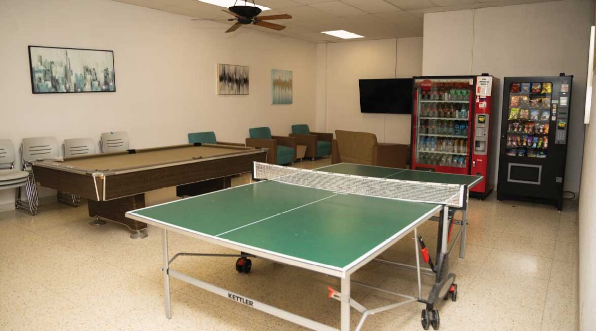 A lounge in Roadman Hall. A pool table and ping pong table are center focus. A mounted TV with seating and 2 vending machines are seen in the background. 