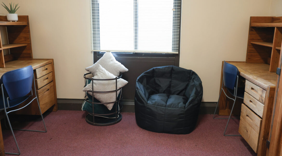 A room in the Plex. Two desks and 2 chair on each side of the wall. A bean bag chair and a basket of pillows sit in the middle of the room.