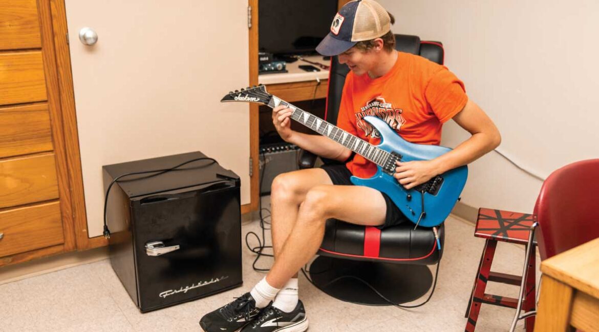 A male student sitting on a chair playing the guitar in a room in Roadman Hall. 
