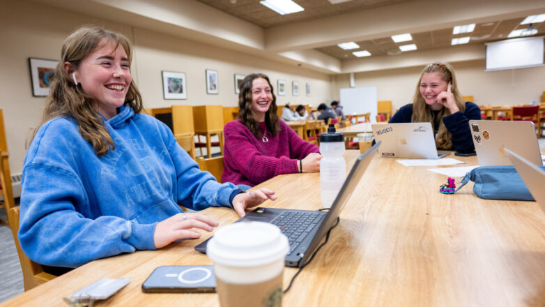 three female students sit around a table in the library laughing while studying